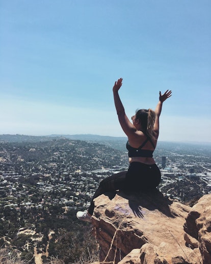 A girl with her back turned to the camera sits on a cliff with her arms raised overlooking a city be...