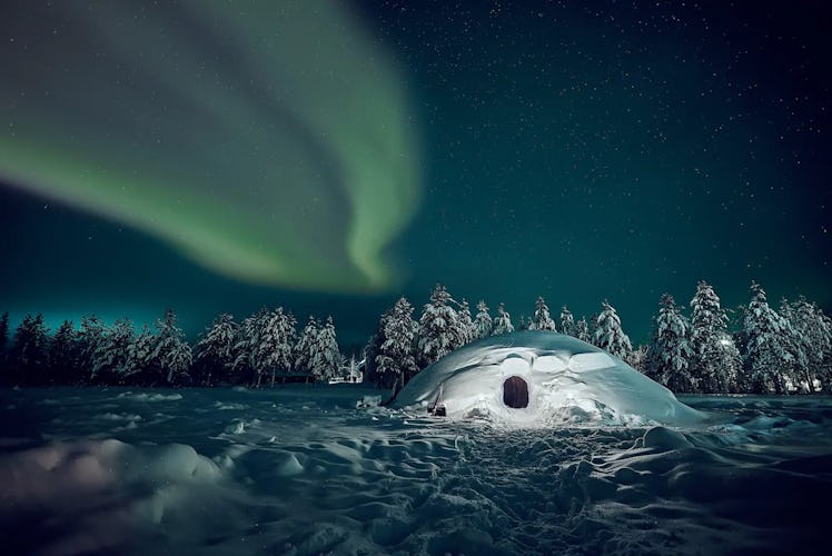 An igloo in the woods at nighttime in Finland has the Northern Lights shining above.