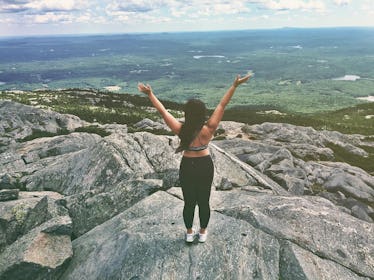 A girl with her back turned to the camera stands at the peak of a mountain looking out over a forest...