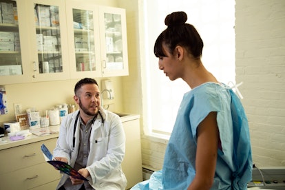 A transgender woman in a hospital gown speaking to her doctor, a transgender man, in an exam room. W...