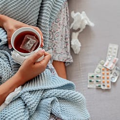A woman with flu sitting covered with blanket with packages of pills in front of her, holding a cup ...