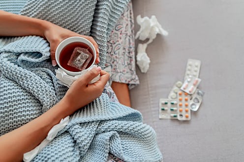 A woman with flu sitting covered with blanket with packages of pills in front of her, holding a cup ...