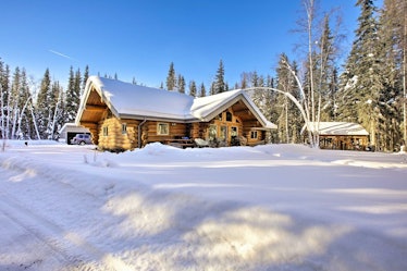 A cozy cabin in North Pole, Alaska is covered in snow and surrounded by tall evergreen trees.
