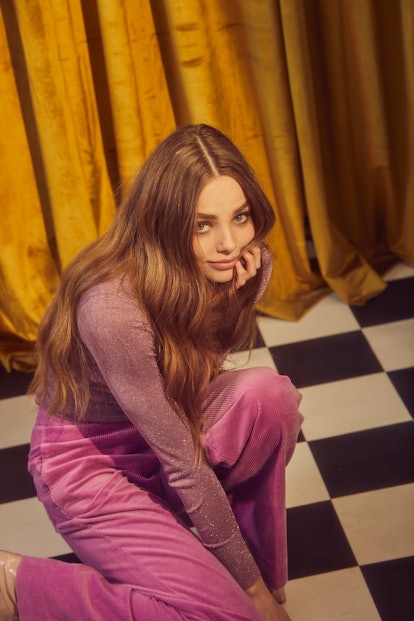 Kristine Froseth in a purple shimmery turtleneck and pink trousers semi-kneeling on the floor and lo...