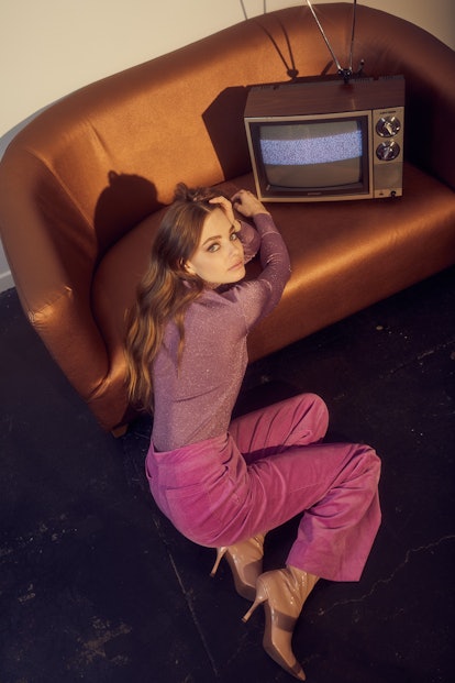 Kristine Froseth in a purple shimmery turtleneck and pink trousers leaning against a bronze couch wi...