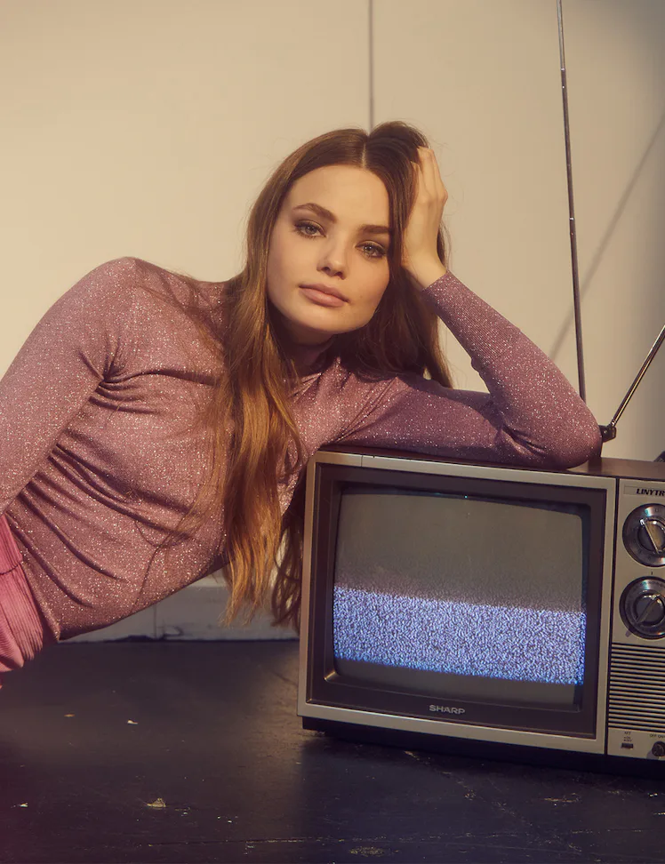 Kristine Froseth in a purple shimmery turtleneck and pink trousers semi-lying and leaning against an...