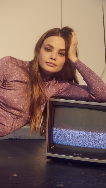 Kristine Froseth in a purple shimmery turtleneck and pink trousers semi-lying and leaning against an...