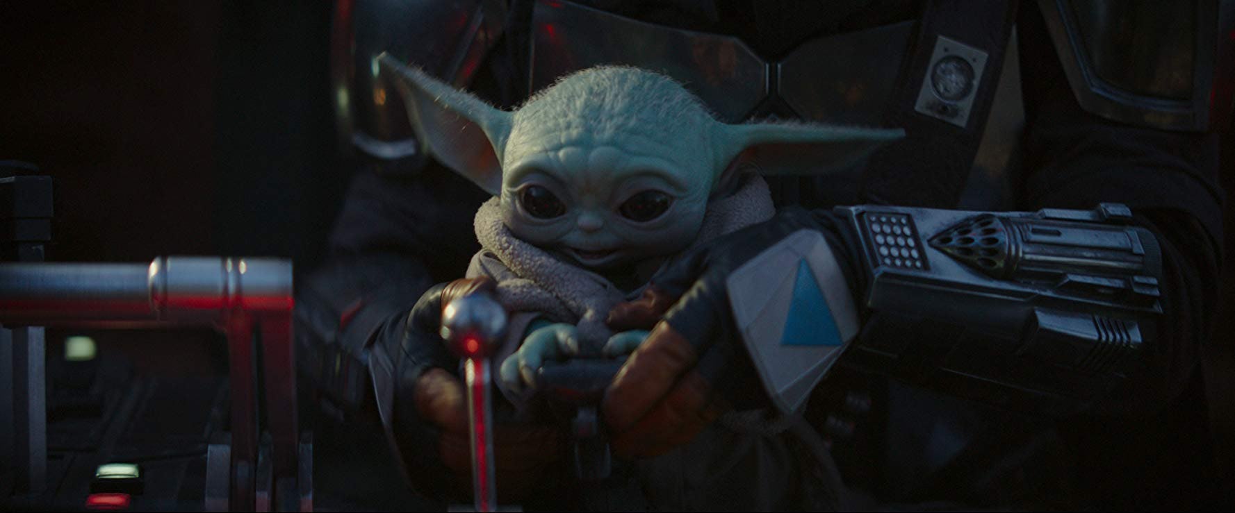 The Mandalorian is holding Baby Yoda, who is smiling and reaching for something. 