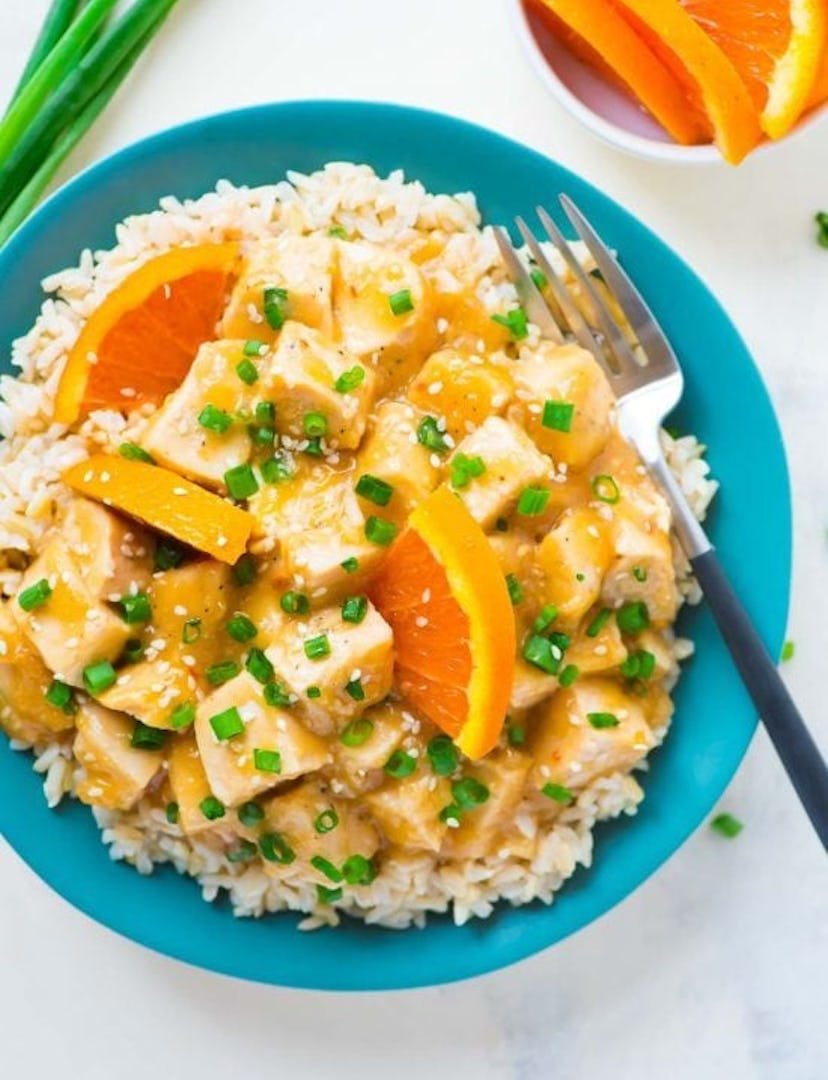 A plate full of orange chicken on white rice garnished with slices of oranges and diced chives