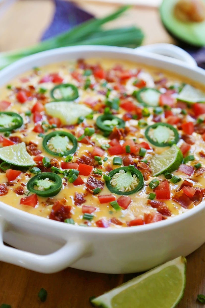 A cheese dip topped with chives, diced tomatoes, bacon bits, limes, and jalapeno peppers