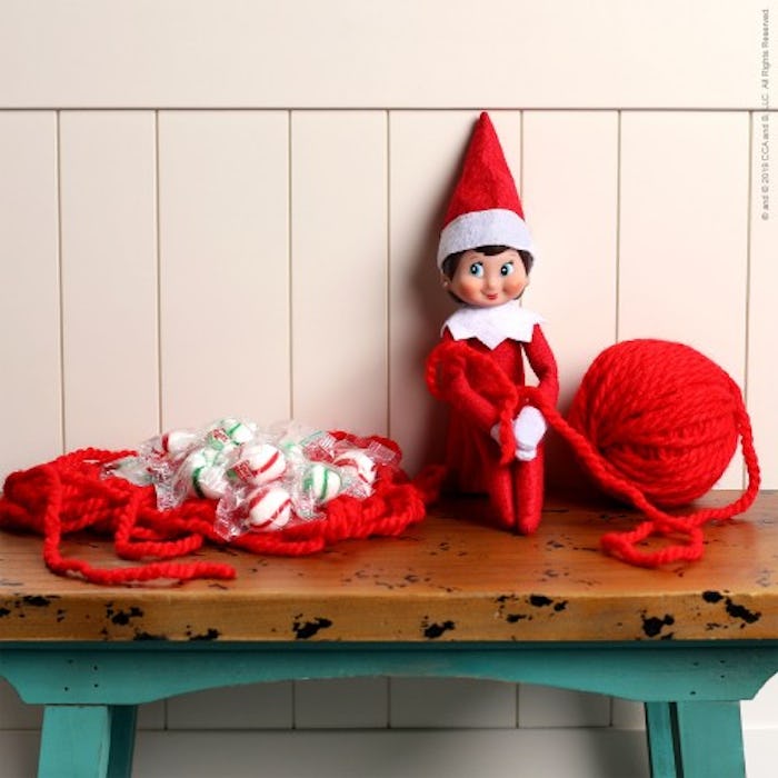 Elf on the shelf sitting on a bench tied up in red yarn with the ball of yarn sitting next to him. 
