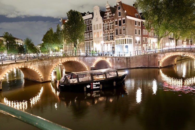 A canal boat rides through the Amsterdam canals during the winter with lights in the background is o...