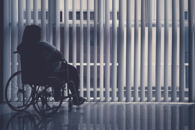 An introverted woman in a wheelchair looking through a window with blinds