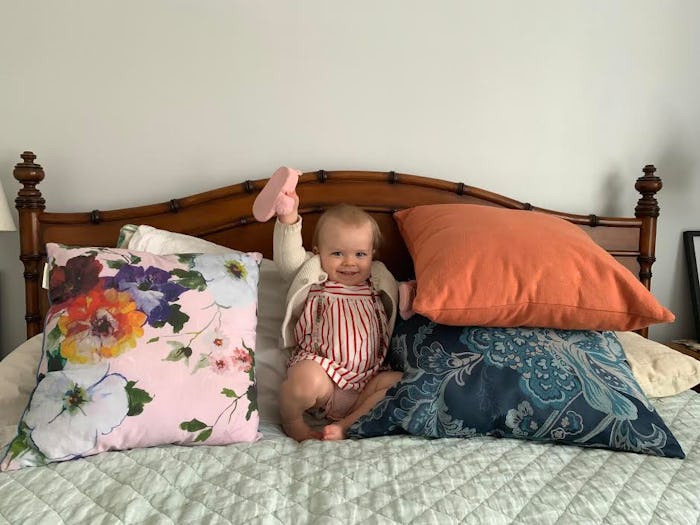 A little girl sitting between big pillows on a bed, holding up a pink slipper