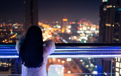 A woman with black long hair, wearing a white shirt looking to a city in front of her from a balcony
