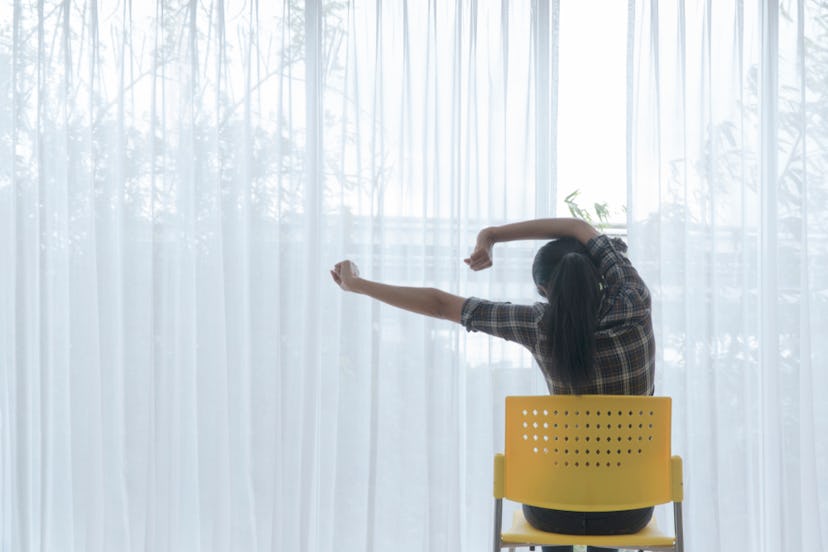 A woman in a tartan shirt stretching her back while sitting in a yellow chair in front of a window.