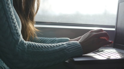 A woman in a mint green cable knit sweater typing on her laptop next to a window.