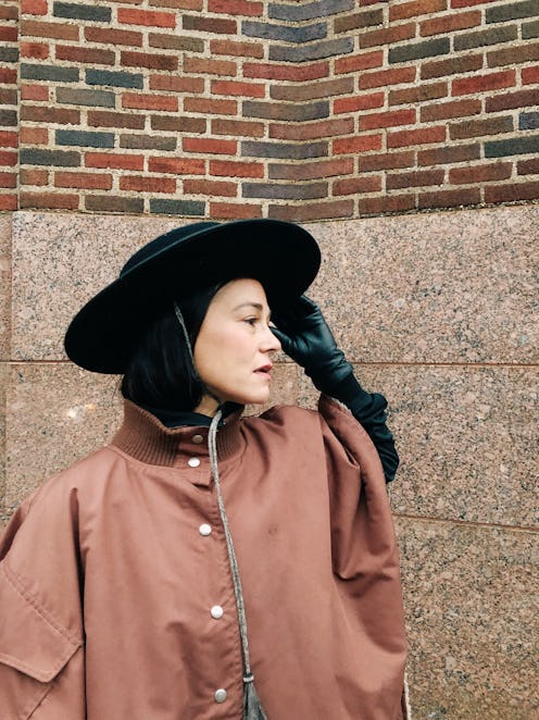 Chef Camille Becerra posing with her hand on her black hat in a tan coat next to a brick wall