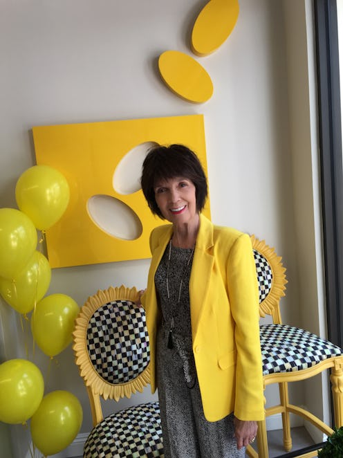 MJ Houghton in a grey pantsuit and a yellow blazer in front of sunflower chekcered chairs and yellow...