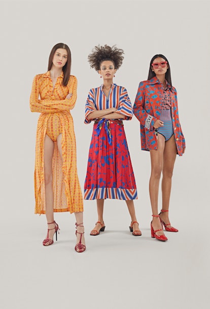 One woman posing in an orange bodysuit, one in a red and blue dress, and the third one in a pink and...