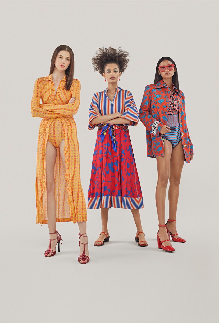 One woman posing in an orange bodysuit, one in a red and blue dress, and the third one in a pink and...