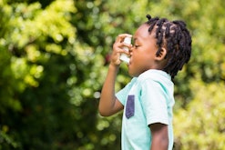A little kid using an inhaler due to asthma, outdoors with trees in the background