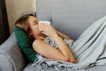 A woman lying on a couch covered with a blanket wiping her nose due to her weakened immune system