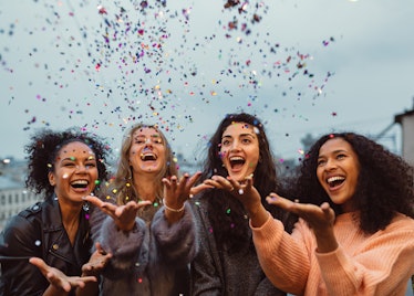 A group of four girls laugh and throw confetti in the air on a rooftop on New Year's Eve.