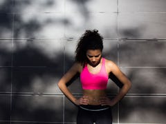 A woman in a pink sports bra and black leggings leaning against a wall as her workout is too intense
