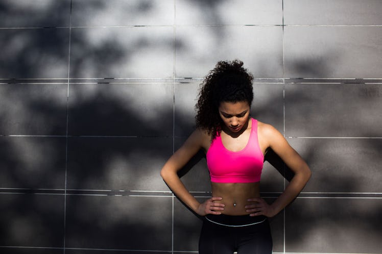 A woman in a pink sports bra and black leggings leaning against a wall as her workout is too intense