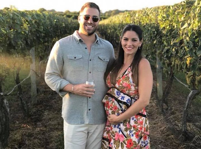 Alyssa Himmel holding her pregnant belly while posing next to her husband in a vineyard