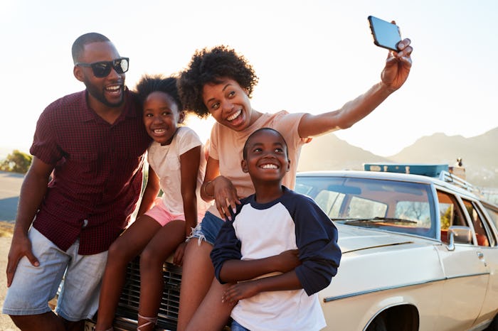A happy family takes a selfie while sitting on the hood of their car in the summer.