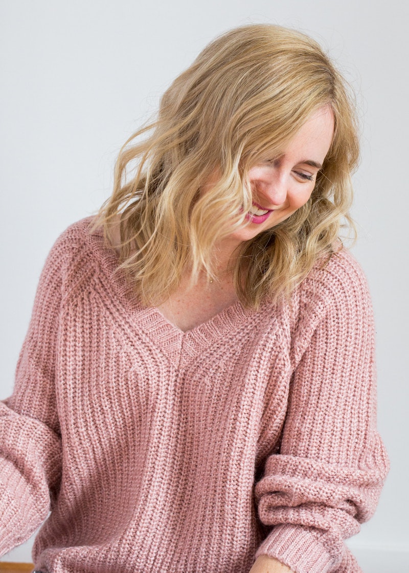 A blonde novelist smiles while wearing a wide pink sweater