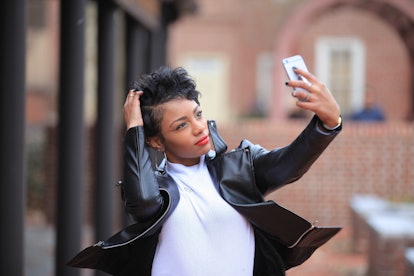 A happy, trendy woman in a leather jacket and turtleneck takes a selfie outside.