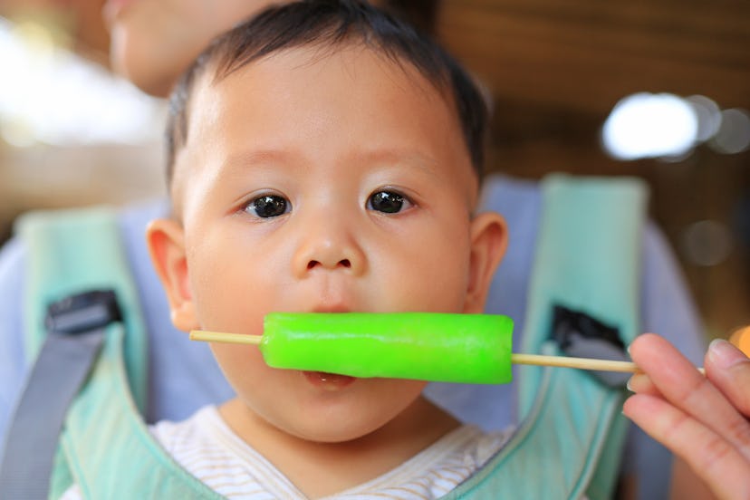 A little baby sitting in a baby carrier worn by his mother and eating a green ice pop