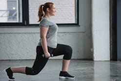 A person with a ponytail and grey t-shirt smiles while performing walking lunges in the gym