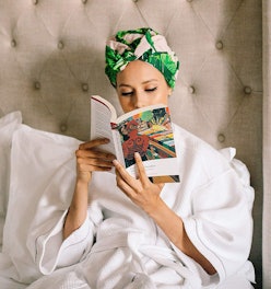 A woman in a white bathrobe and her hair in a colorful sleep cap reading a book while sitting in bed