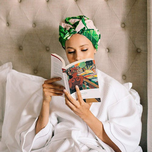 A woman in a white bathrobe and her hair in a colorful sleep cap reading a book while sitting in bed