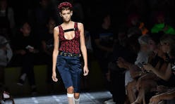 A model walking the runway in a burgundy tank top and denim bermuda shorts 