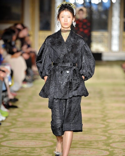 A model walking the runway at a Simone Rocha show in a black wrap jacket and matching bermuda shorts