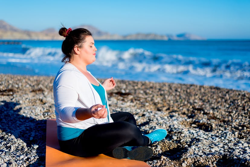 A woman sitting on a yoga mat on the beach meditating while the waves come in