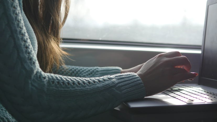 A woman in a mint green sweater typing on her laptop next to a window.