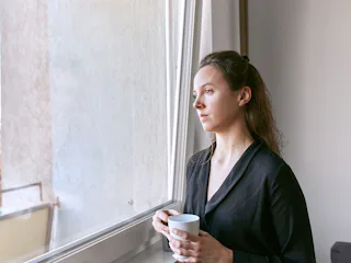 A girl with cold, looking through a window while holding a cup of tea.