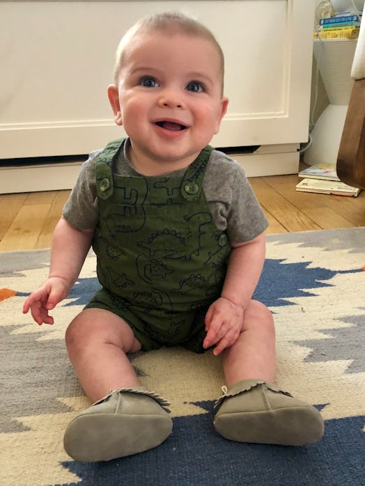 A 6-month-old in a grey tee and dark green overalls sitting on the floor and smiling 