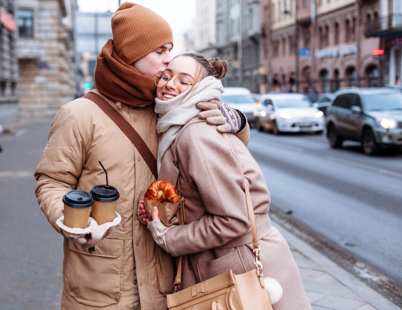 A boy that is holding two coffee to go cups and kissing his girlfriend that is holding a croissant