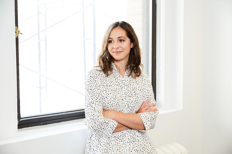 A brunette woman smiling with crossed arms, standing next to a window in a white shirt with black do...