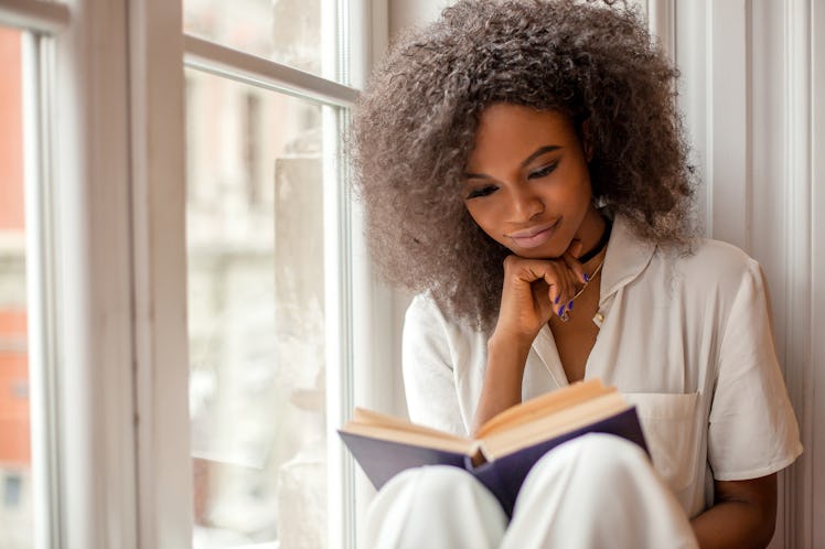 A woman reads a book while sitting next to a window in her pajamas.