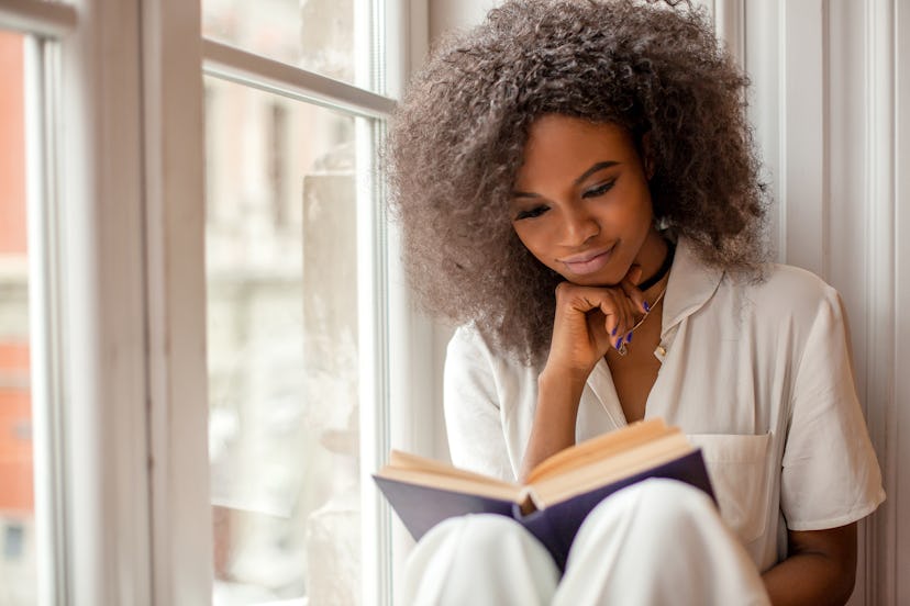 A woman reads a book while sitting next to a window in her pajamas. 