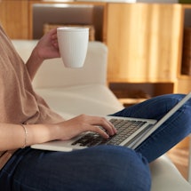 A woman holding a cup in her left hand while typing on her laptop with her right 