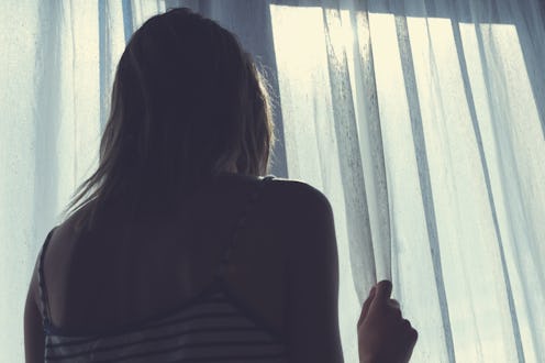 A woman struggling with depression looking through window curtains while standing in a dark room
